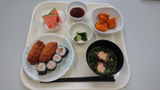 介護施設の食事トレー。唐揚げ、寿司、野菜、汁物が盛られている。